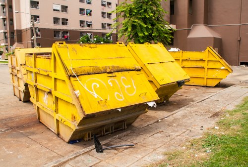 A row of three large, industrial yellow skip bins with metal frames and reinforced sides are placed on a paved sidewalk outside a residential apartment complex. The skip bins have visible weathering and dirt marks, with the front one slightly tilted and marked with white graffiti or signage. They are positioned on a concrete surface adjacent to a patch of grass, with the edge of the pavement and some small debris visible around them. In the background, there are multi-storey residential buildings with brown and beige exteriors, balconies, and green trees providing shade, indicating a typical urban environment in Waterloo. The scene suggests an ongoing rubbish removal or clearance activity, consistent with local office clearance services offered by Office Clearance Waterloo.