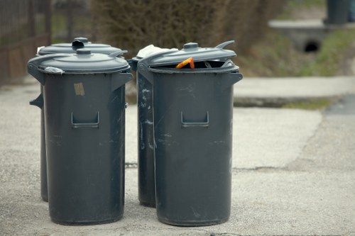 Two large dark grey plastic wheelie bins with closed lids are positioned side by side on a paved pavement surface, likely in an urban or suburban area. The bin on the left has a smooth, matte finish with a horizontal rectangular indentation near the top, while the bin on the right has an open lid revealing discarded waste, including an orange object. The background features a slightly blurred scene with trees and what appears to be a small bridge or culvert, indicating an outdoor setting in or near Waterloo. The image captures the typical appearance of rubbish receptacles used in local waste management, with visible scuff marks and slight wear on the bins, which are standard for handling general household or office waste. This scene relates to rubbish collection and disposal services provided by companies such as Office Clearance Waterloo, serving the Waterloo area postcode, seamlessly fitting into the context of local waste management and disposal activities.