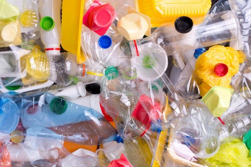 A close-up view of a large pile of discarded plastic bottles, caps, and containers in various colours including clear, yellow, red, green, and blue, with some bottles crumpled or flattened. The plastic waste appears to be gathered on a flat surface, possibly outdoors or on a driveway, in an accumulation that suggests collection or disposal. The textures of the bottles range from smooth and shiny to crinkled, and the different shapes indicate a mixture of beverage bottles, food containers, and caps. The background shows no specific environment but emphasizes the clutter of recyclable plastics, consistent with rubbish removal activities that such companies as Office Clearance Waterloo might manage when clearing waste from residential or commercial premises, particularly in the Waterloo area. The lighting is natural, highlighting the transparency and vibrant colours of the plastic debris, signifying a typical scene of waste ready for collection or transport for recycling or disposal.