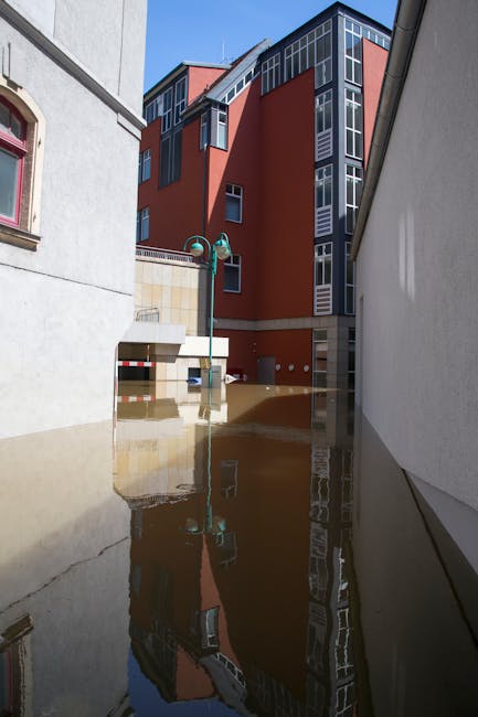 A flooded urban street scene showing several multi-storey buildings with white and pale pink facades in the background. The buildings have large rectangular windows, some with decorative mouldings, an