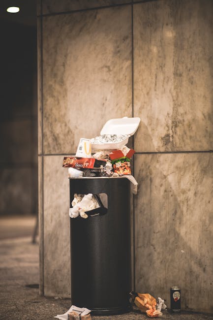 A black wheeled rubbish bin labeled 'St. John's' sits on a sidewalk beside a curb in a residential area at night. The bin is open, filled with various discarded items including cardboard pieces, plast