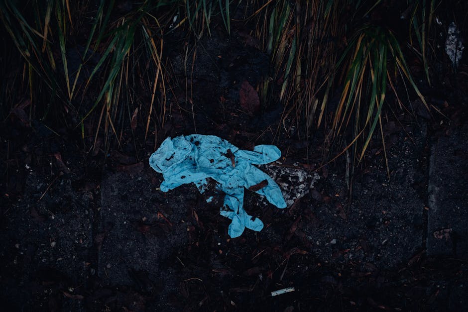 A crumpled, light blue disposable face mask lying on dark, damp soil surrounded by brown leaves and small debris. The mask appears weathered and partially covered in dirt, with elastic ear loops visib