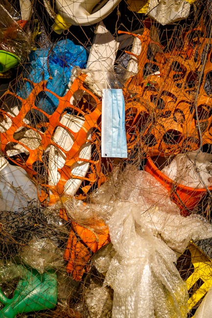 A tangled combination of various waste items contained within an orange plastic barrier, including crumpled clear plastic sheets, a blue disposable face mask hanging loosely amid the clutter, and frag