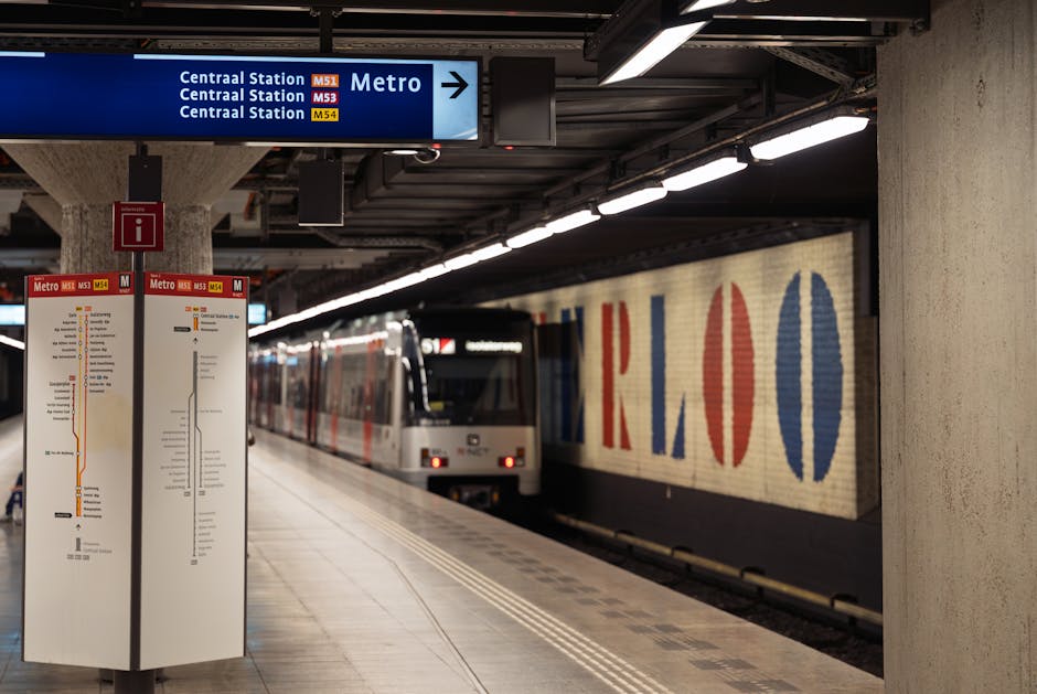 Inside a modern underground railway station, a train with a white body and red and blue detailing is arriving at the platform, which features black and white tiled flooring with tactile paving strips 